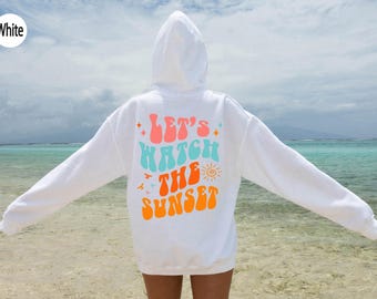 May include: A white hooded sweatshirt featuring a graphic on the back with the text "Let's Watch The Sunset" in pink, light blue, and orange. The design includes small star icons, a sun illustration, and bird silhouettes in a retro font style.