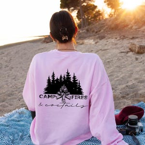 A light pink long-sleeve sweatshirt featuring a black graphic on the back. The design displays a silhouette of pine trees above a campfire with the text CAMP FIRES coctails printed in a combination of bold and script fonts
