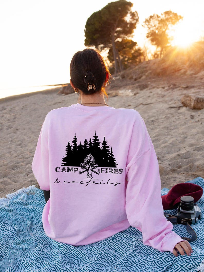 A light pink long-sleeve sweatshirt featuring a black graphic on the back. The design displays a silhouette of pine trees above a campfire with the text CAMP FIRES coctails printed in a combination of bold and script fonts