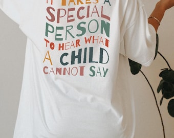 May include: A white t-shirt featuring the text IT TAKES A SPECIAL PERSON TO HEAR WHAT A CHILD CANNOT SAY printed on the back in a multi-colored, wavy font. The lettering uses shades of red, orange, green, and dark blue across the design.