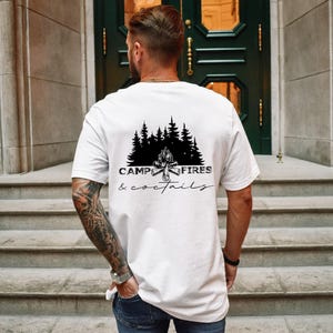 A white short-sleeved t-shirt featuring a black graphic on the back. The design displays a silhouette of pine trees above a campfire, with the text CAMP FIRES COCKTAILS printed in a combination of bold and script lettering