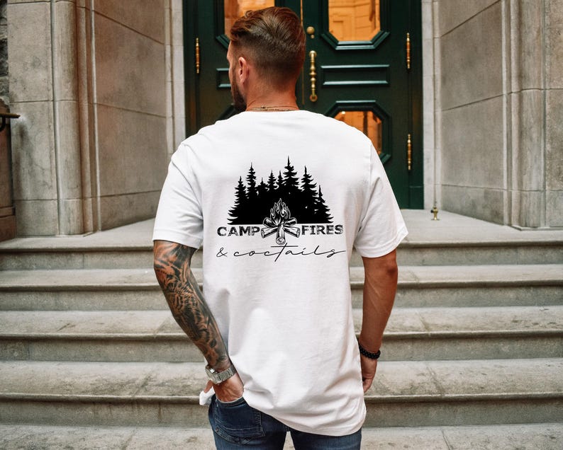A white short-sleeved t-shirt featuring a black graphic on the back. The design displays a silhouette of pine trees above a campfire, with the text CAMP FIRES COCKTAILS printed in a combination of bold and script lettering