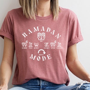 Pu&ograve; includere: Una t-shirt color malva sfumato con una grafica bianca che dice "Ramadan Mode" con una freccia circolare e le parole "Repeat" all'interno della freccia. La grafica include anche le parole "Suhoor, Pray, Quran, Sahur, Iftar, Tarawih" e icone che rappresentano ogni parola.