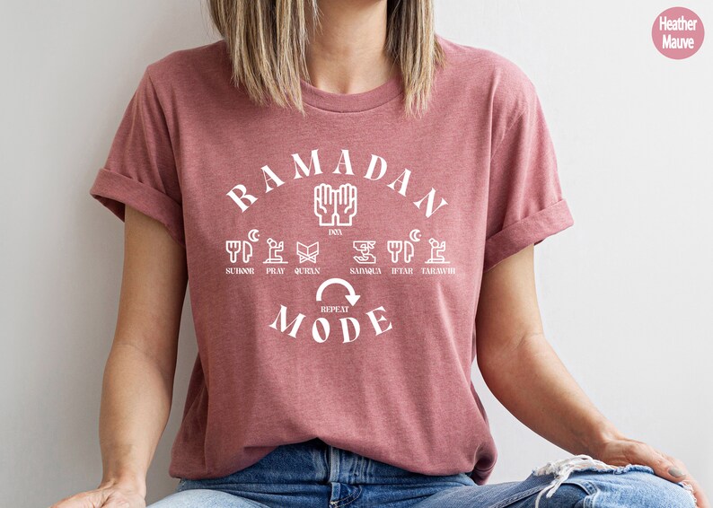 Pu&ograve; includere: Una t-shirt color malva sfumato con una grafica bianca che dice "Ramadan Mode" con una freccia circolare e le parole "Repeat" all'interno della freccia. La grafica include anche le parole "Suhoor, Pray, Quran, Sahur, Iftar, Tarawih" e icone che rappresentano ogni parola.