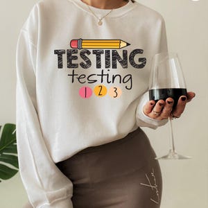 May include: A white long-sleeve sweatshirt featuring a graphic of a yellow pencil above the text "TESTING testing" in black, followed by the numbers "1 2 3" inside pink, yellow, and peach circles. The sweatshirt has a crew neckline.