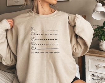 Pode incluir: Uma camisola de gola redonda cor de areia com uma impressão gráfica preta de cinco pequenos animais, cada um seguido por um padrão de linha único, e o texto "WE EACH HAVE OUR OWN TEMPO." A camisola tem mangas compridas e gola e punhos canelados.