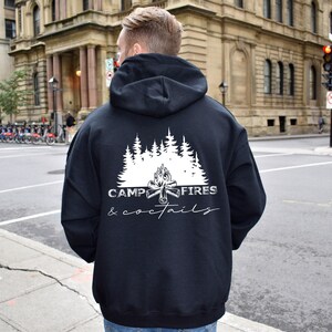 A black hooded sweatshirt featuring a white graphic on the back. The design displays a forest of pine trees above a campfire illustration, with the text CAMP FIRES COCKTAILS printed in a combination of bold and script fonts