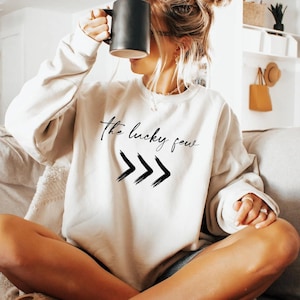 A woman sitting on a couch, holding a coffee mug up to her face, wearing a white sweatshirt with the text "The lucky few" printed on it.