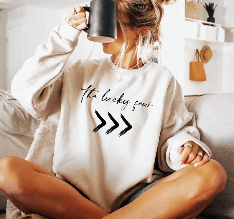 A woman sitting on a couch, holding a coffee mug up to her face, wearing a white sweatshirt with the text "The lucky few" printed on it.