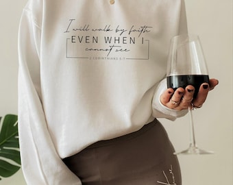 Puede incluir: Una sudadera blanca de manga larga con cuello redondo y texto en negro: I will walk by faith EVEN WHEN I cannot see 2 CORINTHIANS 5:7. La sudadera se combina con leggings marrones y un collar dorado, con una copa de vino en la mano.