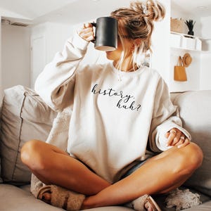 May include: A beige crewneck sweatshirt featuring the black cursive text history. huh? printed on the front. The garment has long sleeves and a relaxed fit, paired with fuzzy beige slide sandals. A solid black mug is held up to the face area.