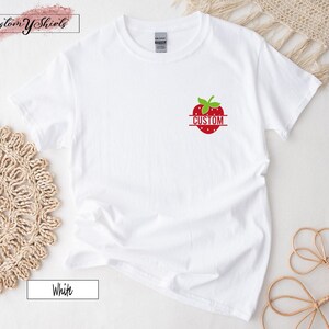 A white short-sleeved t-shirt featuring a small graphic on the left chest area. The graphic depicts a red strawberry with green leaves and the word CUSTOM printed in white capital letters across the center of the fruit