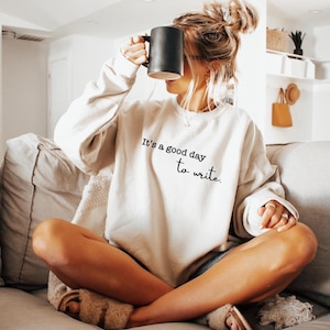 May include: A beige crewneck sweatshirt featuring the black printed text It's a good day to write. The garment has long sleeves and a relaxed fit. A solid black mug is held up near the face, and the outfit is paired with fuzzy open-toe slippers.