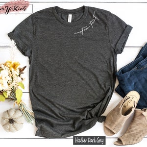 May include: Heather dark grey t-shirt with the text "custom text" written in white on the front. The shirt is laid flat on a white wooden surface with a bouquet of flowers and a pumpkin to the left and a pair of jeans and brown sandals to the right.