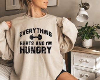 May include: A beige crewneck sweatshirt featuring black distressed text that reads EVERYTHING HURTS AND I'M HUNGRY with a graphic of a dumbbell positioned between the words EVERYTHING and HURTS, printed on the center of the chest.