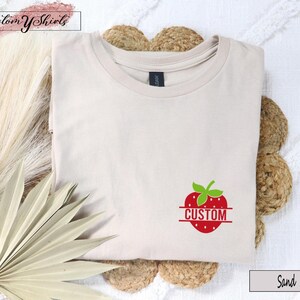 A sand-colored crew neck t-shirt featuring a red strawberry graphic on the lower right side. The strawberry has a green stem and a horizontal line across the center with the word CUSTOM printed in red capital letters