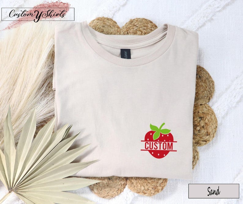 A sand-colored crew neck t-shirt featuring a red strawberry graphic on the lower right side. The strawberry has a green stem and a horizontal line across the center with the word CUSTOM printed in red capital letters