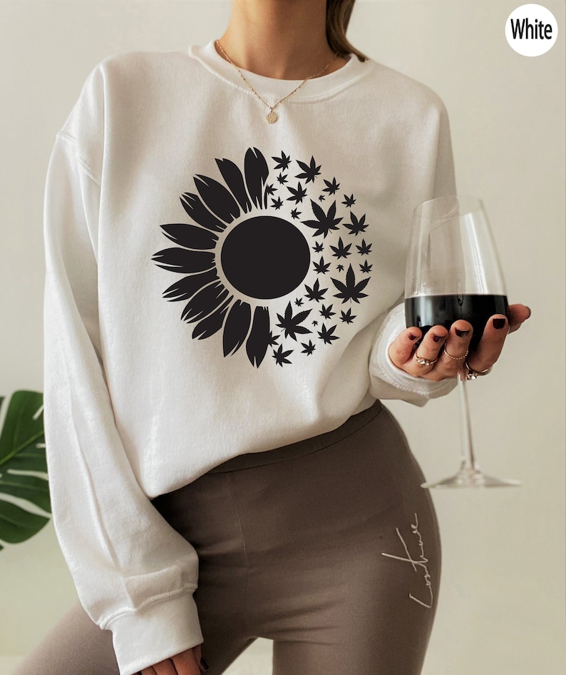 May include: White sweatshirt with a black sunflower and marijuana leaf design. The sweatshirt is being worn by a person holding a glass of red wine.