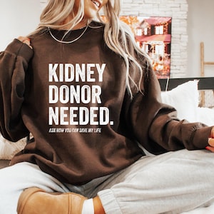 May include: A dark brown long-sleeve sweatshirt featuring the white text KIDNEY DONOR NEEDED. and the smaller text ASK HOW YOU CAN SAVE MY LIFE printed on the front. The garment has a crew neckline and a relaxed fit with ribbed cuffs.