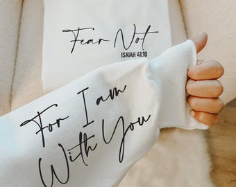 Puede incluir: Una sudadera blanca de manga larga con texto negro en cursiva en el pecho que dice Fear Not ISAIAH 41:10. La manga muestra texto negro adicional en cursiva que dice For I am With You, escrito con una fuente fluida.
