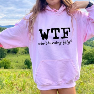 May include: Pink hoodie with a black graphic that says "WTF who's turning fifty?"
