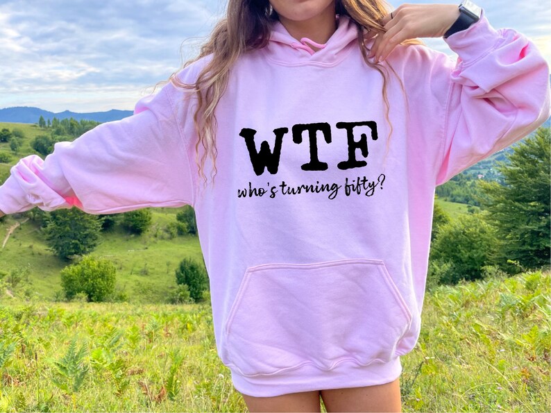 May include: Pink hoodie with a black graphic that says "WTF who's turning fifty?"