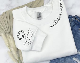 May include: A white crewneck sweatshirt featuring black cursive text that reads autism mom near the neckline. The sleeve displays a black outline of a single puzzle piece above the cursive text custom name. The garment has a standard collar.
