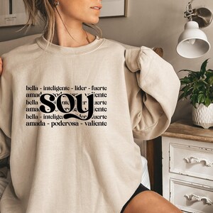May include: A beige crewneck sweatshirt featuring black text printed on the front. The design includes the word "soy" in a large, bold font centered over repeating lines of Spanish words including "bella," "inteligente," "lider," "fuerte," "amada," "poderosa," and "valiente."