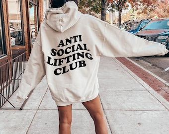 May include: A cream-colored hooded sweatshirt featuring the black text ANTI SOCIAL LIFTING CLUB printed on the back. The garment has long sleeves, a hood, and a relaxed fit, showcasing a bold, arched typography design across the upper back area.