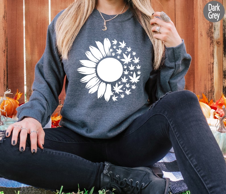 May include: A dark gray sweatshirt with a white graphic of a sunflower with marijuana leaves. The sweatshirt is being worn by a person sitting on a blanket.