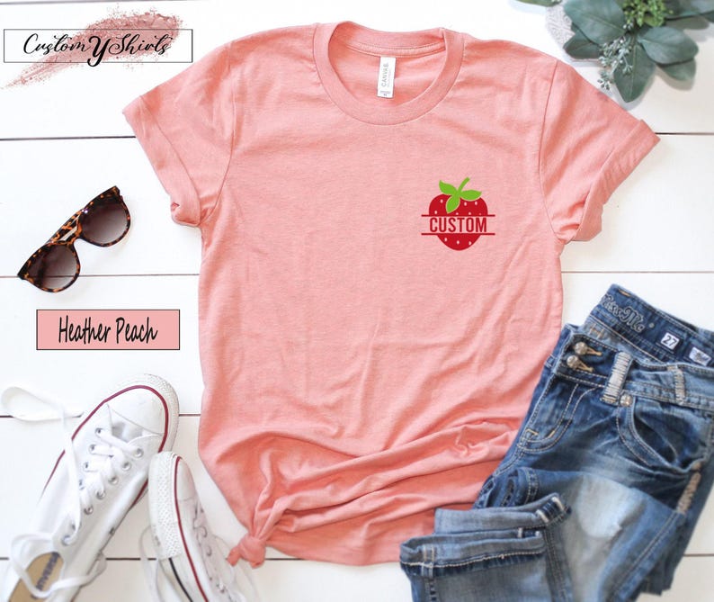 A heather peach short-sleeve t-shirt featuring a small graphic on the left chest of a red strawberry with green leaves and the word CUSTOM printed in white across the center of the fruit