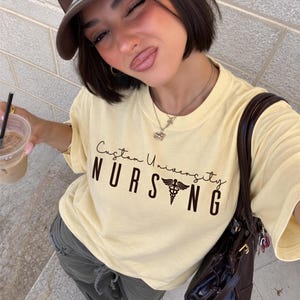 May include: A pale yellow short-sleeve t-shirt featuring the text Custom University NURSING in dark brown, with a caduceus symbol replacing the letter I. The shirt is paired with olive green cargo pants and a brown baseball cap.