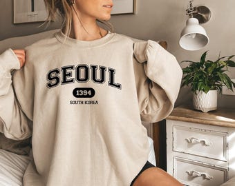 May include: A beige crewneck sweatshirt featuring black collegiate-style lettering that reads SEOUL, with the number 1394 inside a black oval and the text SOUTH KOREA printed below in a smaller, simple black font on the chest area.