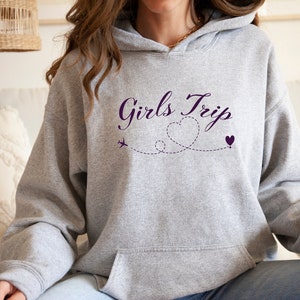 K&ouml;nnte beinhalten: Grauer Kapuzenpullover mit dem Text "Girls Trip" in lilafarbener Kursivschrift. Der Text wird von einem lilafarbenen Flugzeug und zwei lilafarbenen Herzen begleitet, die durch eine gepunktete Linie verbunden sind.