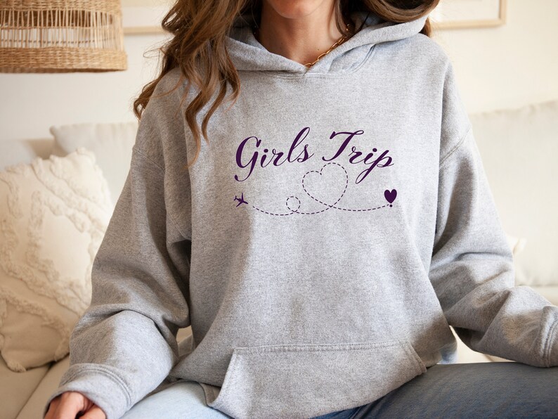 K&ouml;nnte beinhalten: Grauer Kapuzenpullover mit dem Text "Girls Trip" in lilafarbener Kursivschrift. Der Text wird von einem lilafarbenen Flugzeug und zwei lilafarbenen Herzen begleitet, die durch eine gepunktete Linie verbunden sind.