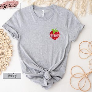 A sport grey short-sleeve t-shirt featuring a small graphic on the left chest area. The graphic depicts a red strawberry with green leaves and a horizontal line across the center containing the word CUSTOM in red capital letters