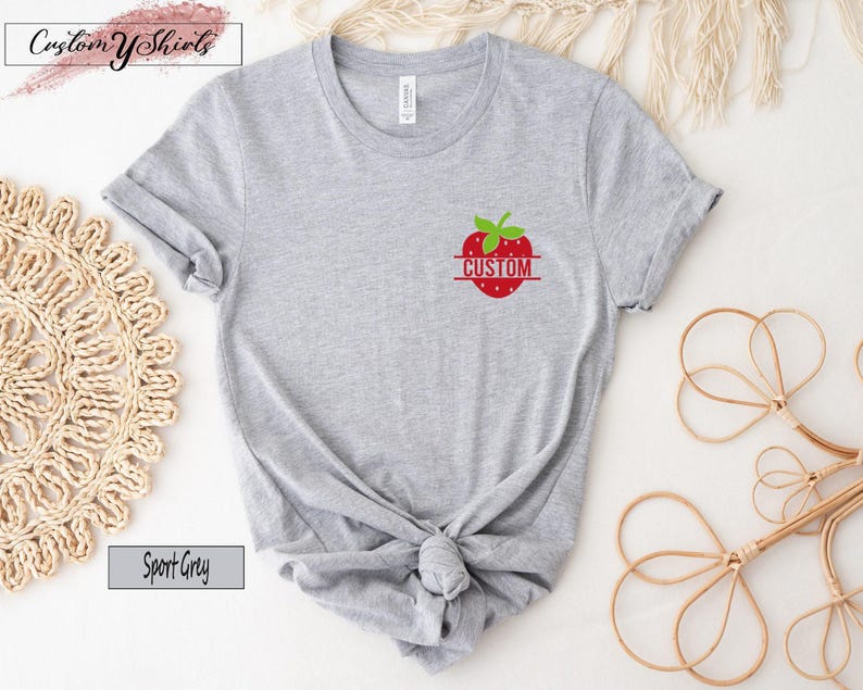 A sport grey short-sleeve t-shirt featuring a small graphic on the left chest area. The graphic depicts a red strawberry with green leaves and a horizontal line across the center containing the word CUSTOM in red capital letters