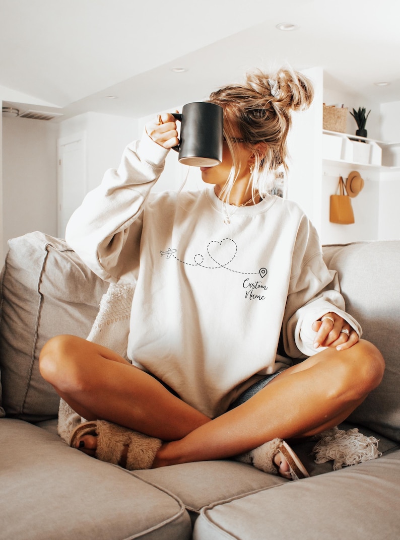 May include: A white sweatshirt with a black airplane, heart, and pin design. The text "Custom Name" is printed below the design.