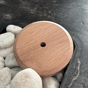 Puede incluir: Un disco redondo de madera con un agujero en el centro. El disco es de color marrón claro y tiene una superficie lisa. Está sentado sobre un lecho de piedras blancas.