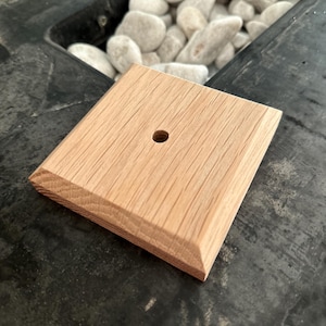 Puede incluir: Un bloque de madera cuadrado, de color claro, con un borde biselado y un agujero centrado. La veta de la madera es visible y el bloque parece nuevo. El artículo es probablemente un componente para un proyecto más grande o un elemento decorativo.