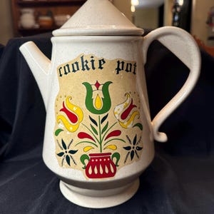 May include: A white ceramic cookie jar shaped like a teapot with a speckled glaze. The jar has a floral design with red, yellow, green, and black accents. The jar has the words "cookie pot" written on the front.