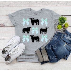 May include: A gray t-shirt with a design of black cows and green bows. The shirt is laid out on a wooden surface with a pair of white sneakers and a pair of blue jeans.
