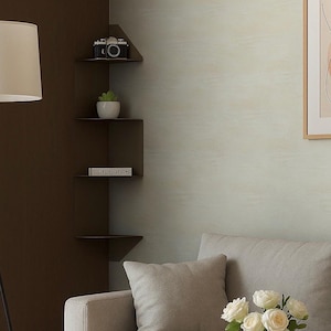 Puede incluir: Una esquina de salón moderna con una estantería esquinera de metal marrón oscuro. Las estanterías exhiben una cámara vintage, una pequeña suculenta en maceta y un libro. La habitación tiene una paleta de colores neutros con un sofá gris y un jarrón de rosas blancas.