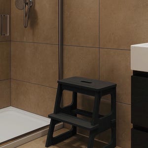 May include: A dark grey wooden step stool with two steps. The stool is in a bathroom setting, with a shower and a vanity visible in the background. The step stool is designed to provide assistance in reaching higher places.