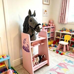 May include: A pink wooden hobby horse stable stand featuring a purple unicorn graphic on the side and the text Hobby Horse Stables. A grey plush hobby horse with a black bridle is mounted on top, and four small toy horses stand on the bottom shelf.