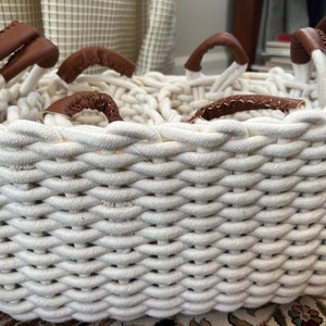 Peut inclure: Un panier en corde blanche avec quatre poignées en cuir marron. Le panier est tissé avec un motif serré et complexe.