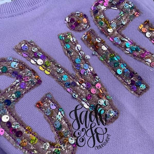 Puede incluir: Una sudadera de color morado claro con letras grandes de estilo bloque decoradas con lentejuelas iridiscentes multicolores. Las letras están delineadas con costuras de color morado oscuro sobre una base de tela texturizada marrón claro. Un logotipo negro es visible.