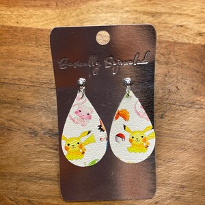 Puede incluir: Un par de pendientes blancos en forma de l&aacute;grima con un Pikachu amarillo y otros personajes de Pok&eacute;mon impresos en ellos. Los pendientes est&aacute;n en una tarjeta plateada con el texto "Beautifully Blinged" impreso en ella.
