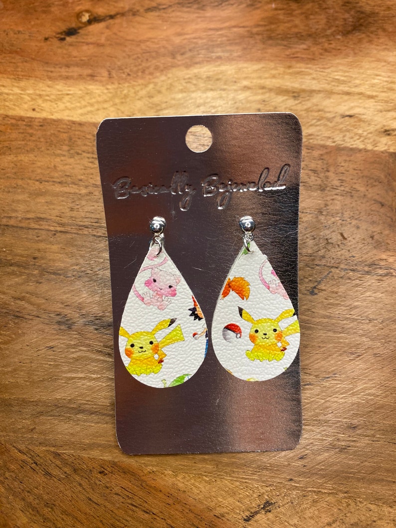 Puede incluir: Un par de pendientes blancos en forma de l&aacute;grima con un Pikachu amarillo y otros personajes de Pok&eacute;mon impresos en ellos. Los pendientes est&aacute;n en una tarjeta plateada con el texto "Beautifully Blinged" impreso en ella.