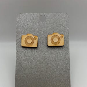 May include: Two wooden camera-shaped stud earrings. The earrings are light brown with a detailed camera design. They are displayed on a gray card with a hole at the top. The earrings are a photography-themed accessory.